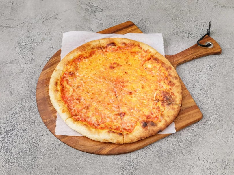 A whole cheese pizza on a wooden serving board, placed on a gray textured surface.
