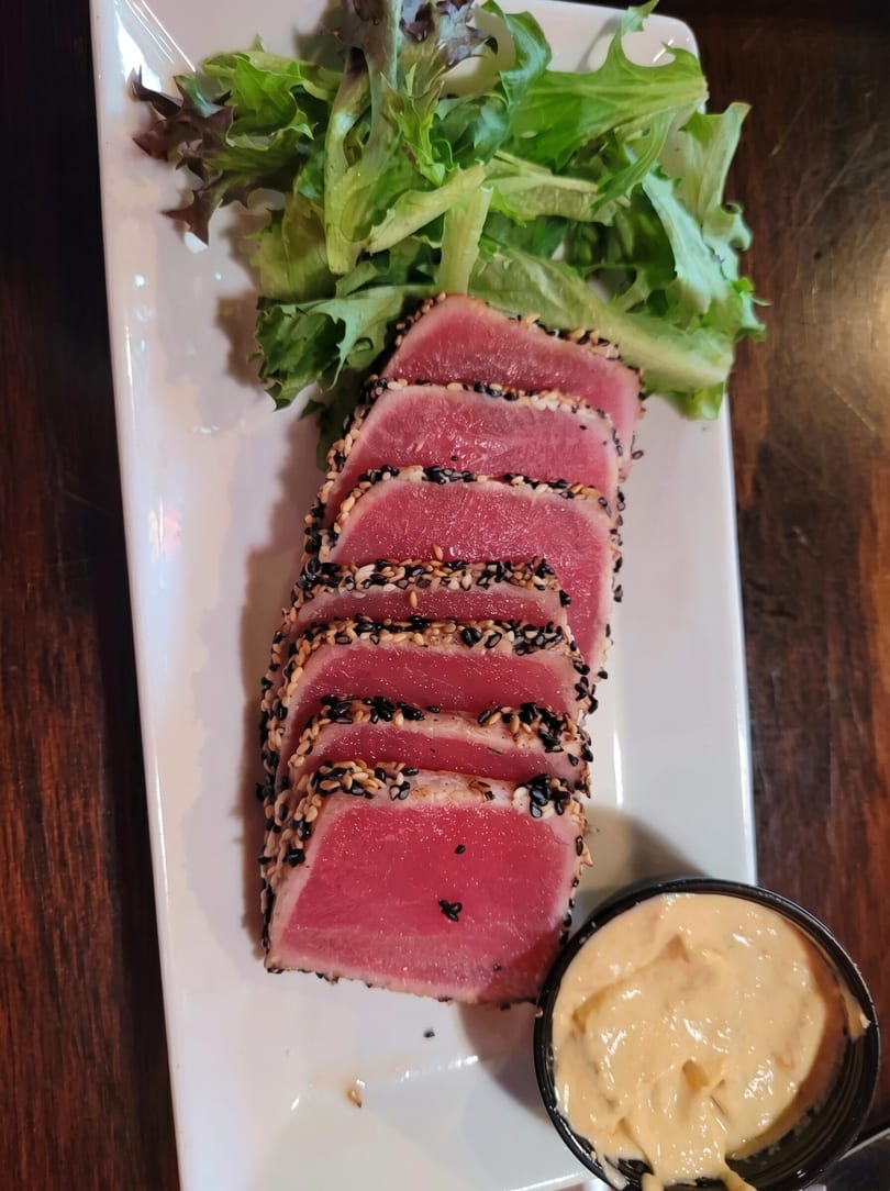 Seared tuna slices with sesame crust, fresh greens, and a side of creamy sauce on a white plate.